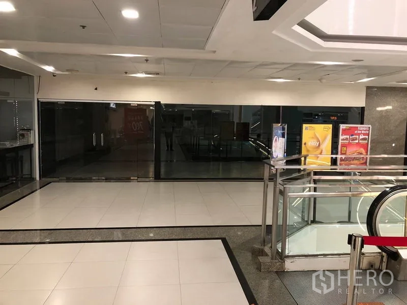 Retail Space for rent in Khlong Toei, Bangkok - Empty retail unit with wide glass storefront beside an escalator inside a Bangkok shopping mall