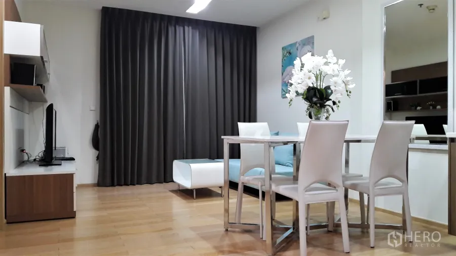 Condominium for rent in Khlong Toei, Bangkok - Living–dining space with wood floors, TV console, sofa, large curtains and a modern dining set with flowers.