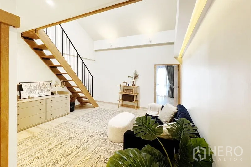 Townhouse for rent in Khlong Toei, Bangkok - High-ceiling family room with mezzanine staircase, sofa seating and minimalist decor.
