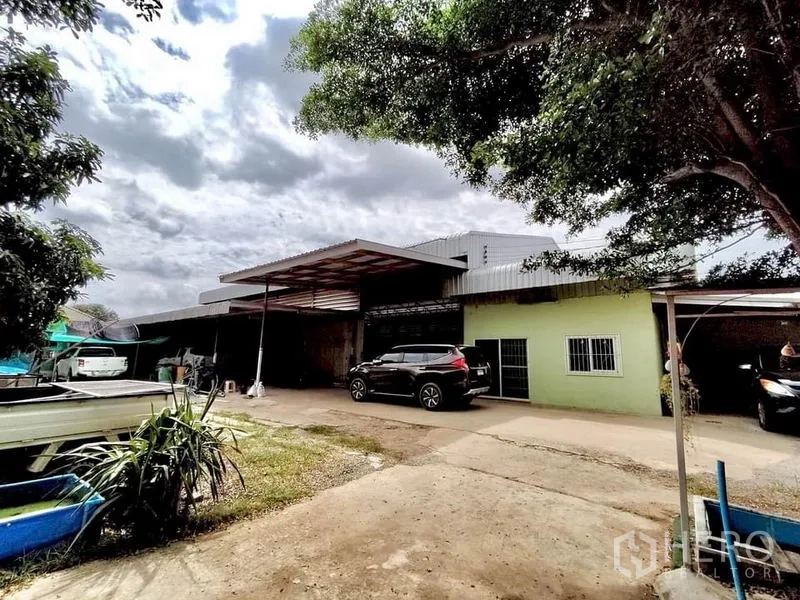 Factory for sale in Lat Lum Kaeo, Pathum Thani - Front view of single-storey factory with covered loading bay, concrete yard and parked vehicles shaded by trees.