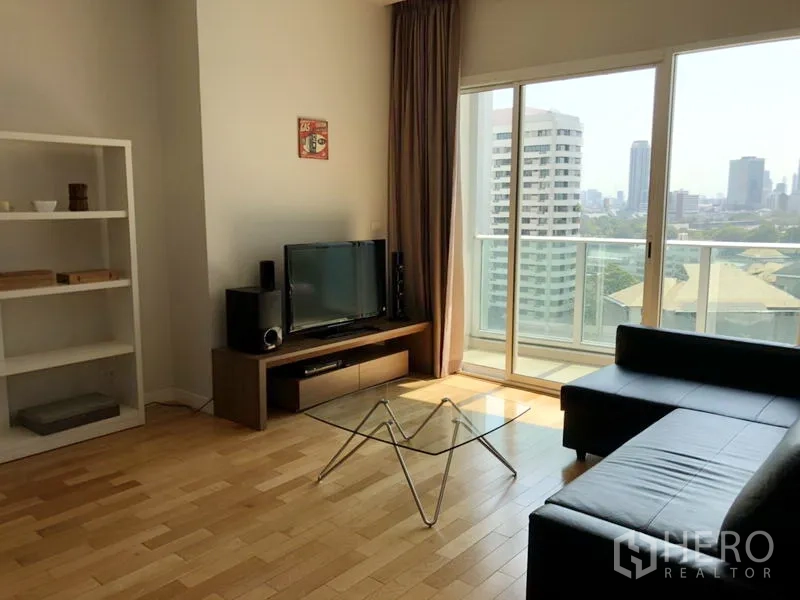 Condominium for rent in Khlong Toei, Bangkok - Bright living room with black leather sofa, glass coffee table, TV console and sliding doors to a balcony with skyline view.