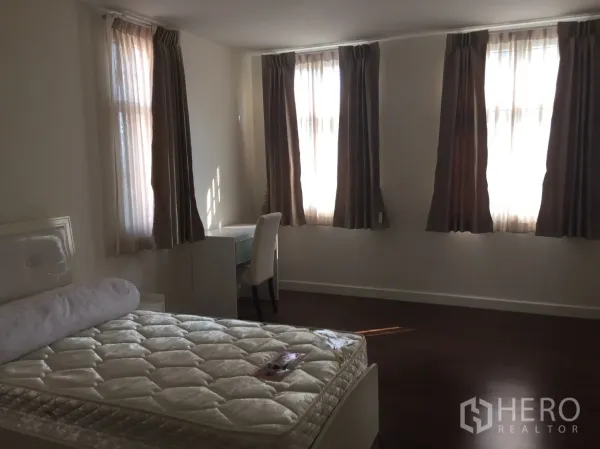Townhouse for rent in Watthana, Bangkok - Spacious bedroom with multiple curtained windows, bed, and study desk on wooden flooring.