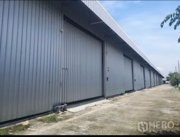 Warehouse for rent in Lam Luk Ka, Pathum Thani - Row of metal-clad warehouse units with large sliding doors and a wide concrete driveway.