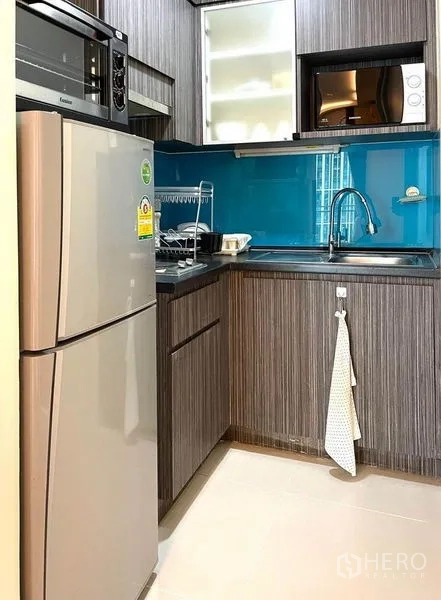 Condominium for rent in Huai Khwang, Bangkok - Compact L-shaped kitchen with wood cabinets, blue glass backsplash and appliances.