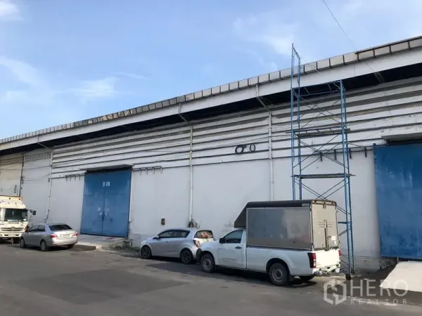货栈 出租在Bang Phli, Samut Prakan - 仓库外立面，蓝色钢质装卸门，路边通道与停放的车辆。