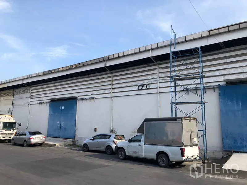 Warehouse for rent in Bang Phli, Samut Prakan - Long single-storey warehouse exterior with blue steel loading doors, roadside access and parked delivery vehicles.
