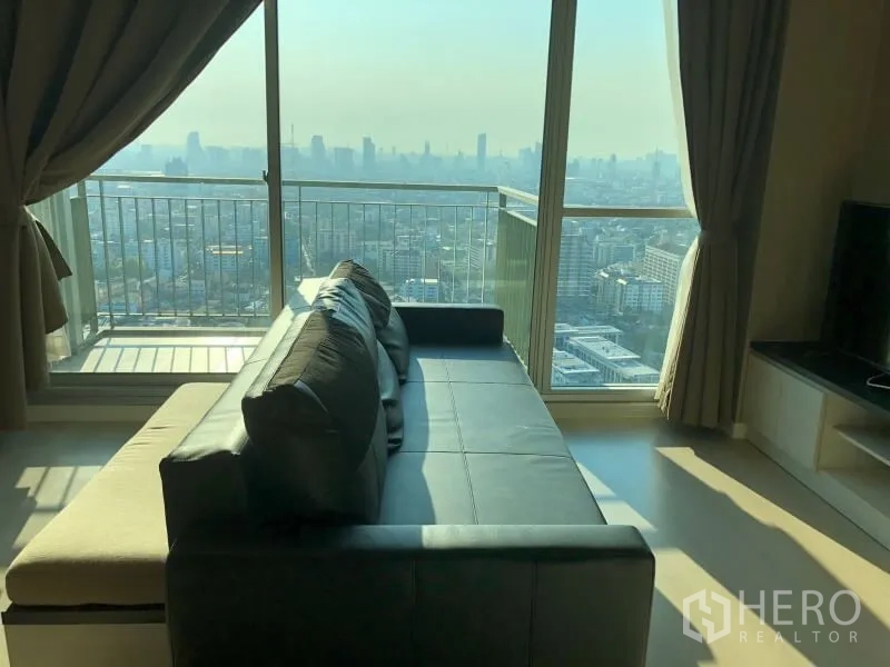 Condominium for rent or sale in Huai Khwang, Bangkok - Living room with sectional sofa opening to a balcony with sweeping city views.