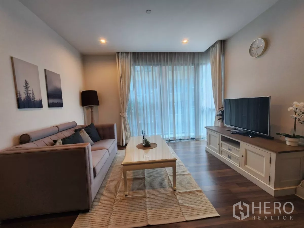 Condominium for rent in Phra Khanong, Bangkok - TV corner opposite a sofa in an airy living room with curtains and wood flooring.
