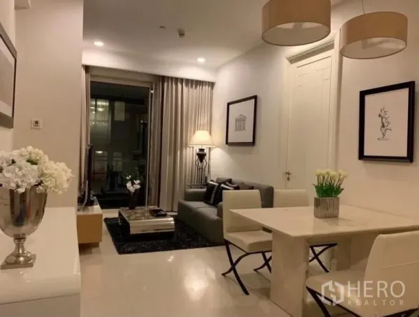 Condominium for rent in Pathum Wan, Bangkok - Open-plan living and dining area with stone dining table, pendant lights, gray sofa, framed art, and sliding glass door to a balcony at night.