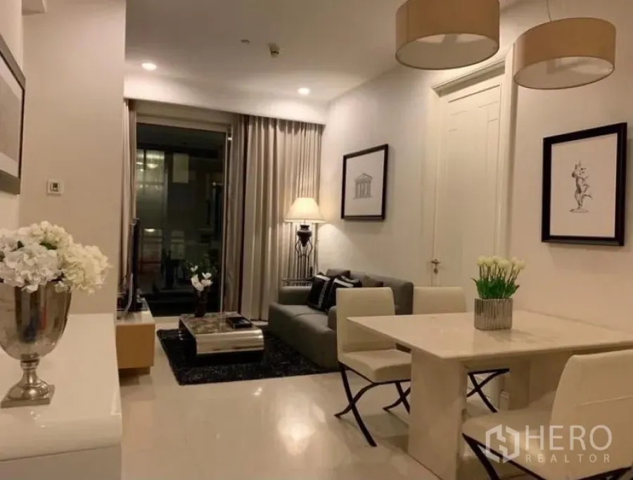 Condominium for rent in Pathum Wan, Bangkok - Open-plan living and dining area with stone dining table, pendant lights, gray sofa, framed art, and sliding glass door to a balcony at night.