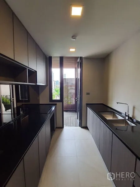 Condominium for sale in Huai Khwang, Bangkok - Kitchen view toward the service balcony with built-in cabinets and twin-basin sink under recessed lights.