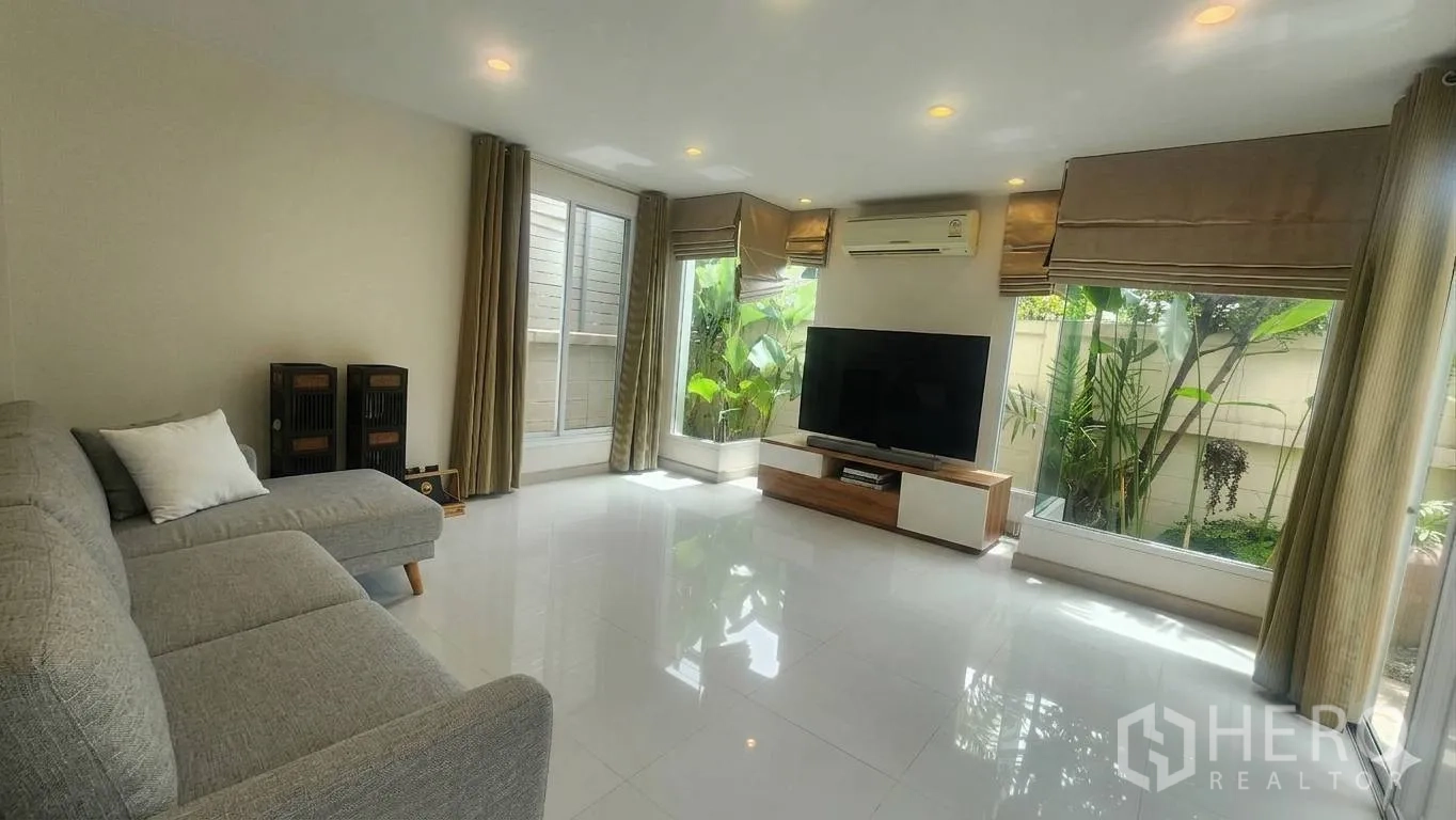 Detached House for rent in Watthana, Bangkok - Bright living room with large windows, gray sectional sofa, and TV unit.