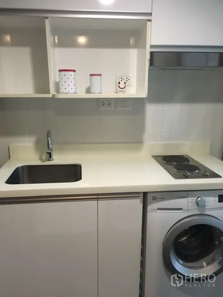 Condominium for rent in Khlong Toei, Bangkok - Compact white kitchen with sink, electric cooktop and under-counter washing machine.