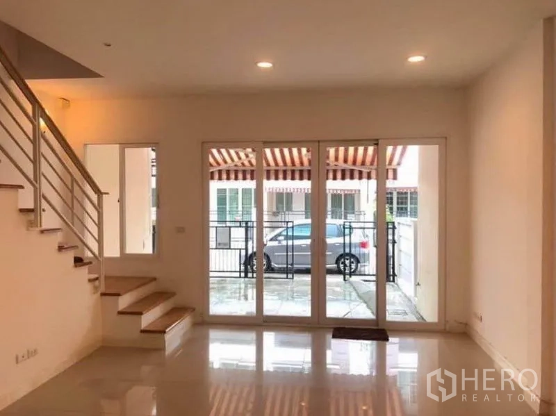 联排别墅 出租在Wang Thonglang, Bangkok - 通透客厅通过大幅玻璃门连接前庭与停车位，可见金属栅栏与遮阳篷。