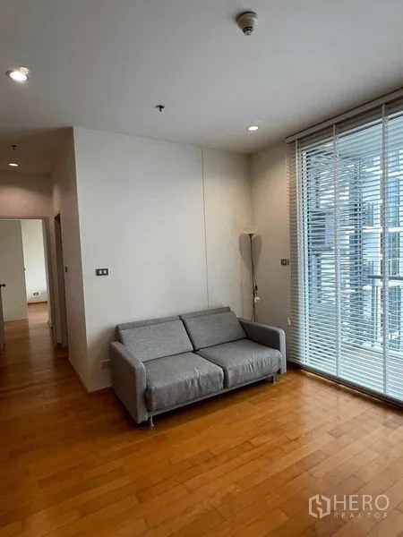 Condominium for rent in Ratchathewi, Bangkok - Living area with gray sofa beside floor-to-ceiling windows and blinds.