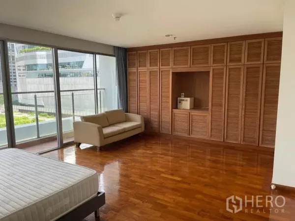 公寓 出租在Pathum Wan, Bangkok - 宽敞卧室，落地窗与木质柜墙，带沙发和可通往阳台的城市景观。