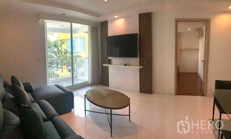 Condominium for rent or sale in Khlong Toei, Bangkok - Living room with TV wall, access to the balcony, and doorway to a bedroom.