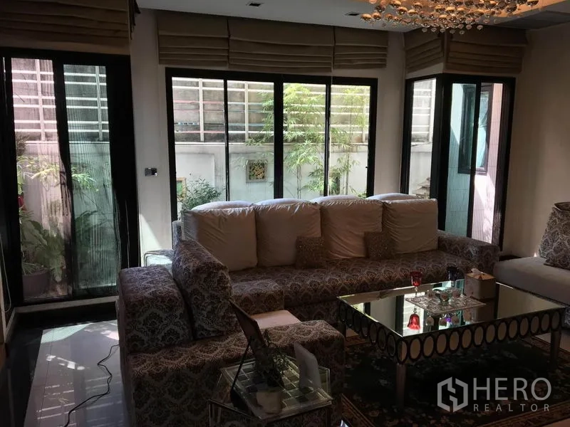 บ้าน ให้เช่าในKhlong Toei, Bangkok - อีกมุมของห้องนั่งเล่น โซฟาเข้ามุม โต๊ะกลางกระจกเงา และประตูกระจกบานเลื่อนมองเห็นสวน