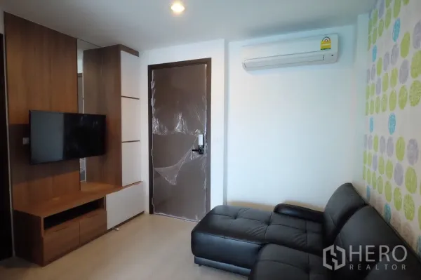 Condominium for rent in Sathon, Bangkok - TV wall and entry door in the living room with black leather sofa and air conditioner at a Sathon condo.