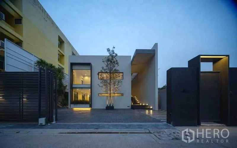 带办公室的家 出售在Wang Thonglang, Bangkok - 面向街道的现代立面，带大门与宽阔铺装停车庭院，暮色氛围。