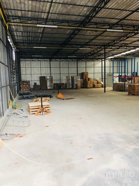 Warehouse for rent in Wang Thonglang, Bangkok - Spacious warehouse interior with concrete floor, steel truss roof, and stacked pallets and boxes along the walls.