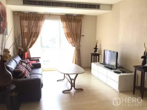公寓 出租在Watthana, Bangkok - 明亮客厅，皮沙发、电视柜与茶几，落地推拉门通向阳台并配窗帘。
