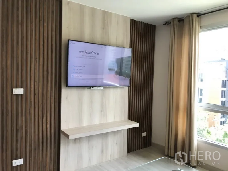 Condominium for rent in Watthana, Bangkok - Sunny room with TV mounted on a wood accent wall beside a large window and beige curtains.