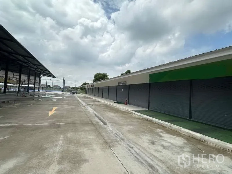 Warehouse for rent in Sai Noi, Nonthaburi - Long row of shuttered units beside a wide driveway and loading lane.