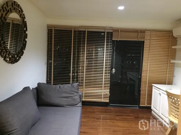 公寓 出租在Watthana, Bangkok - 书房/起居角落，灰色沙发、圆形装饰镜、内置书桌柜体与落地百叶帘。