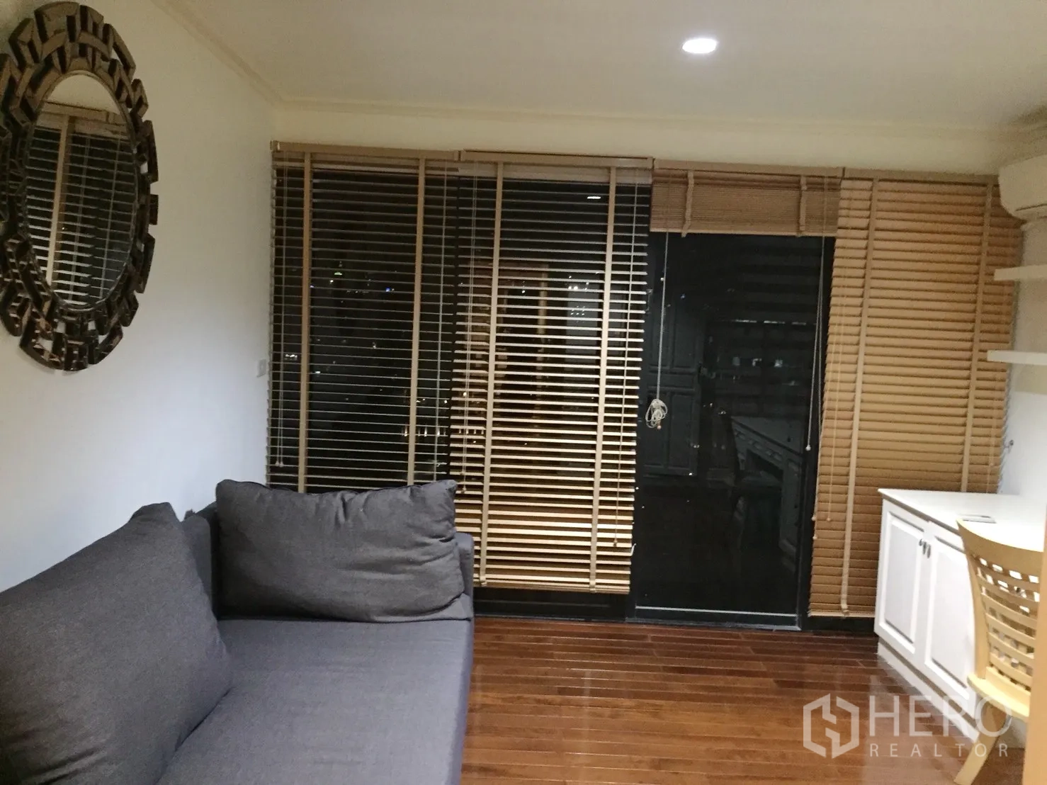 公寓 出租在Watthana, Bangkok - 书房/起居角落，灰色沙发、圆形装饰镜、内置书桌柜体与落地百叶帘。