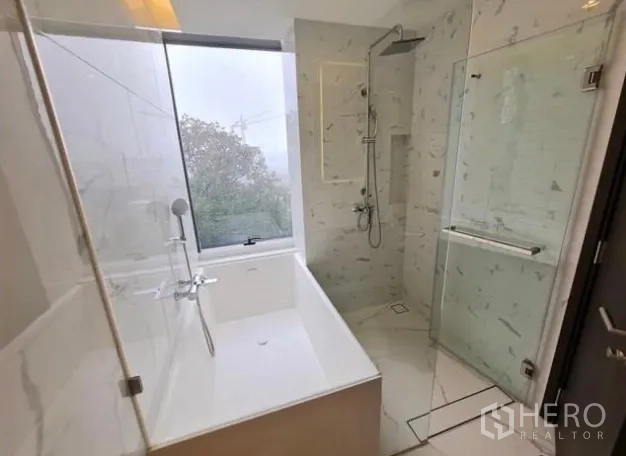 Condominium for rent in Khlong Toei, Bangkok - Bright bathroom with large window, separate bathtub and glass‑enclosed rain shower in marble finish.