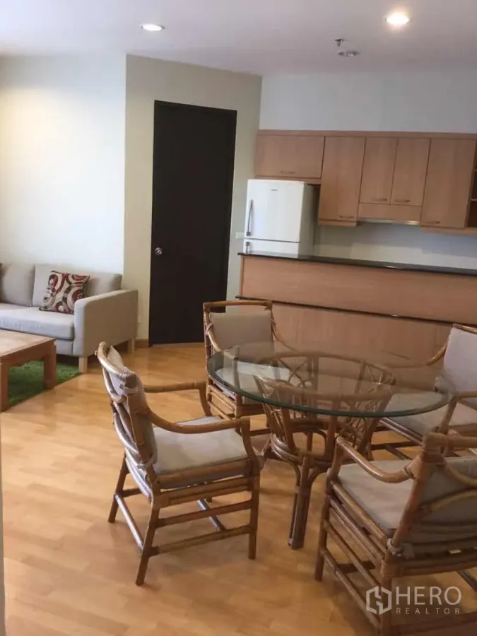 Condominium for rent in Khlong Toei, Bangkok - Open-plan living and dining area with rattan chairs, glass table, sofa and a kitchenette with wood cabinets.