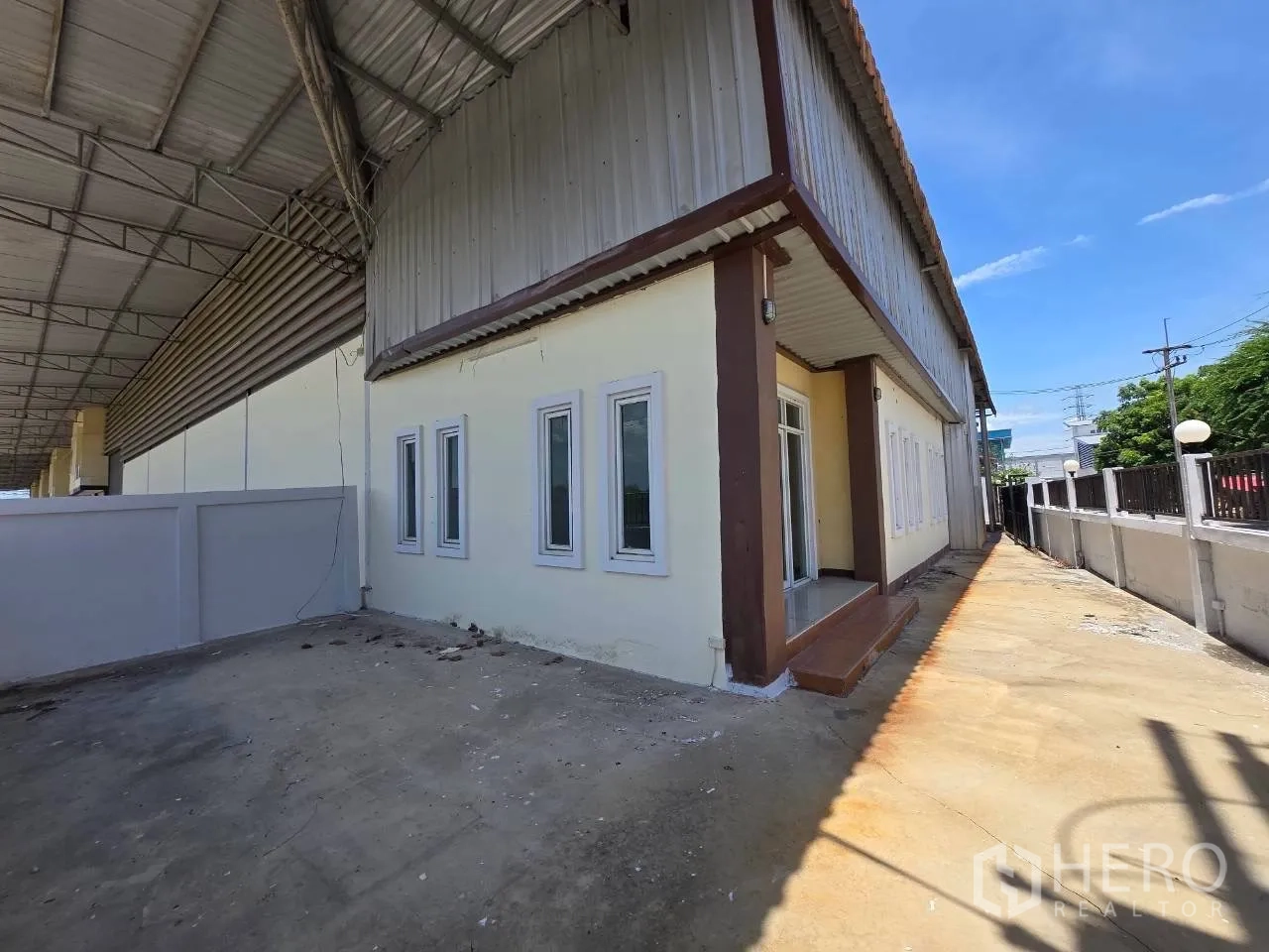 Warehouse for rent or sale in Mueang Samut Sakhon, Samut Sakhon - Corner view of the warehouse exterior with entrance steps, windows, and a concrete driveway under a large roof canopy.