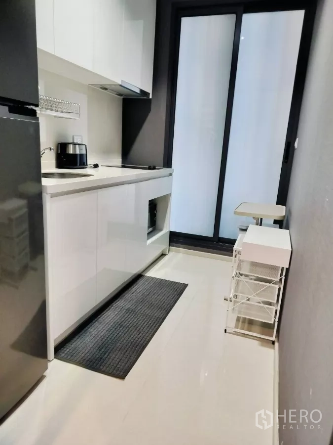 Condominium for rent in Khlong Toei, Bangkok - Compact galley kitchen with white cabinets, sink, hob and sliding glass door.