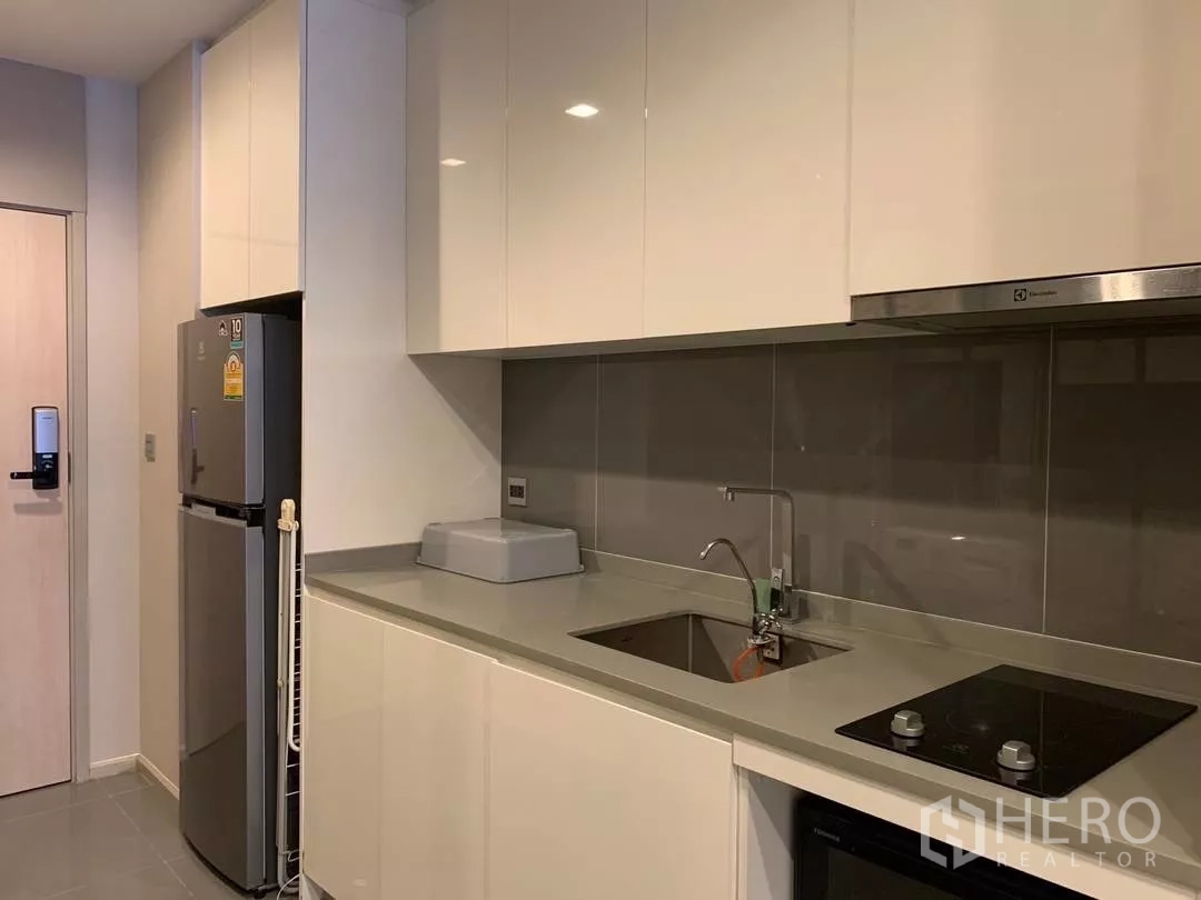 Condominium for rent or sale in Watthana, Bangkok - Modern galley kitchen with glossy cabinets, electric cooktop, hood, sink and full-size refrigerator.