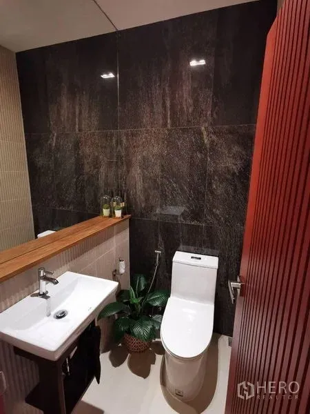Townhouse for rent or sale in Yan Nawa, Bangkok - Contemporary powder room with dark stone feature wall, white sink, toilet, wooden ledge and indoor plant.