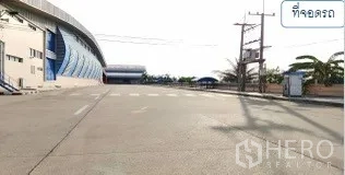 Warehouse for rent in Mueang Pathum Thani, Pathum Thani - Wide concrete driveway in front of the warehouse with a guard booth and landscaping.