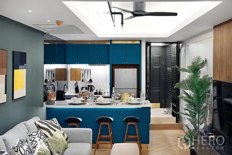 Condominium for rent in Bang Rak, Bangkok - Open-plan condo kitchen with teal cabinets, breakfast island set for dining, and a cozy living area beneath a ceiling fan.