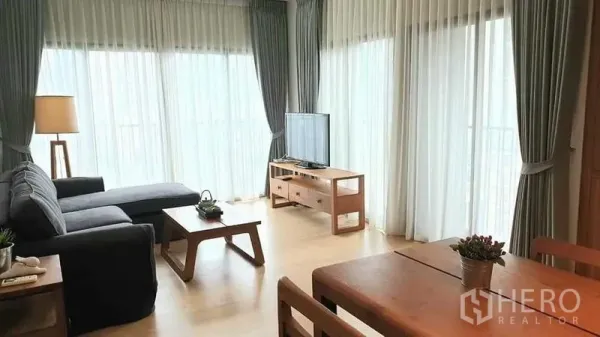 คอนโด ให้เช่าหรือขายในWatthana, Bangkok - ห้องนั่งเล่นมุม โซฟา L ชั้นวางทีวี และหน้าต่างสูงจรดเพดานพร้อมม่านโปร่ง
