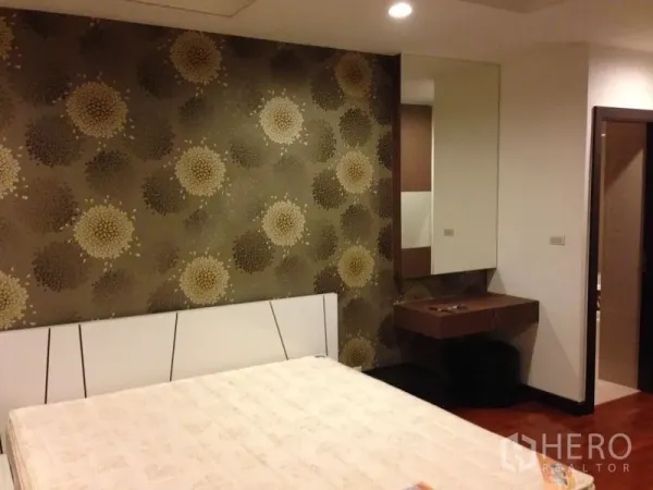 คอนโด ให้เช่าในWatthana, Bangkok - ห้องนอนหลักมีวอลเปเปอร์ลายดอกไม้ หัวเตียงบุนวม และโต๊ะเครื่องแป้งติดกระจก ข้างประตูห้องน้ำ