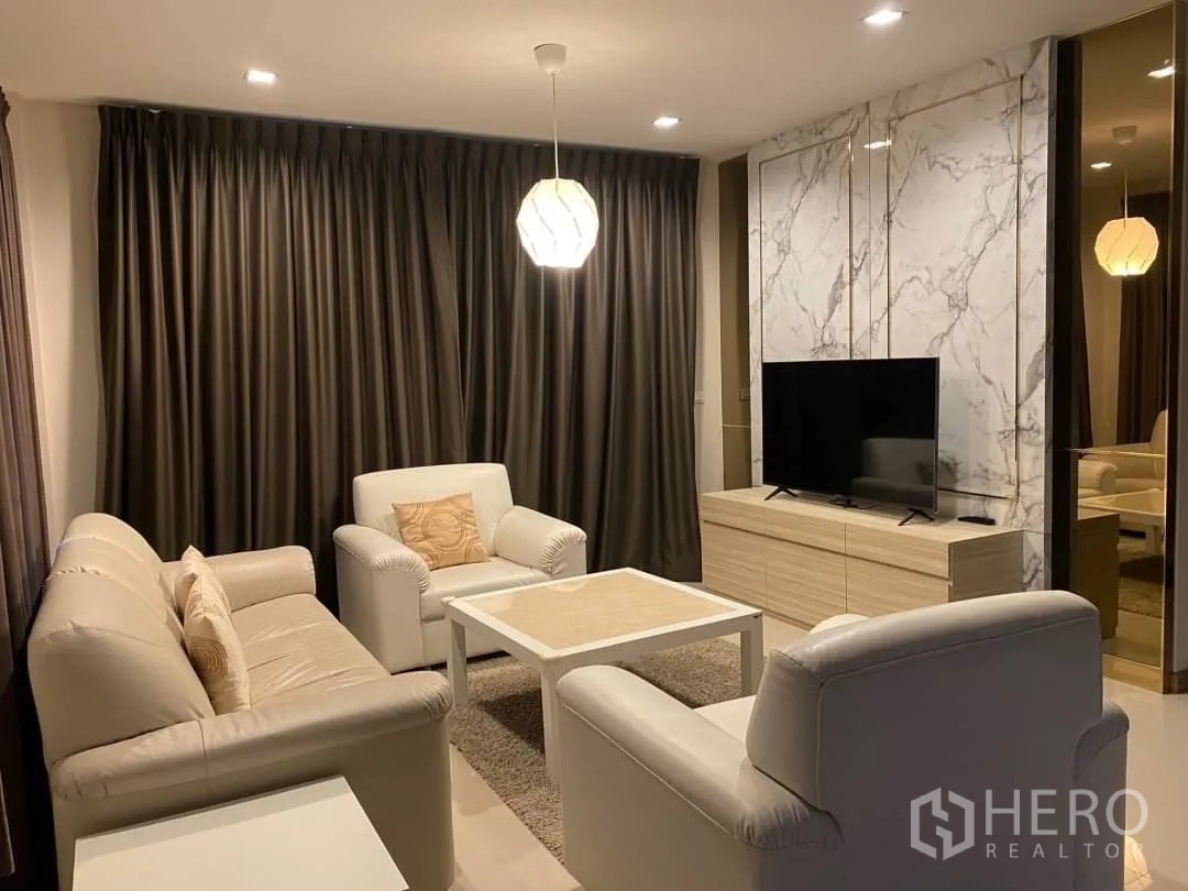 Detached House for rent or sale in Prawet, Bangkok - Cozy living room with cream sofas, a TV and a marble accent wall with dark drapes.