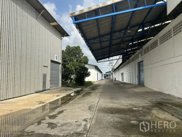 Factory for rent in Nonthaburi - Roller shutter bays along a concrete lane between warehouses with roof overhang.