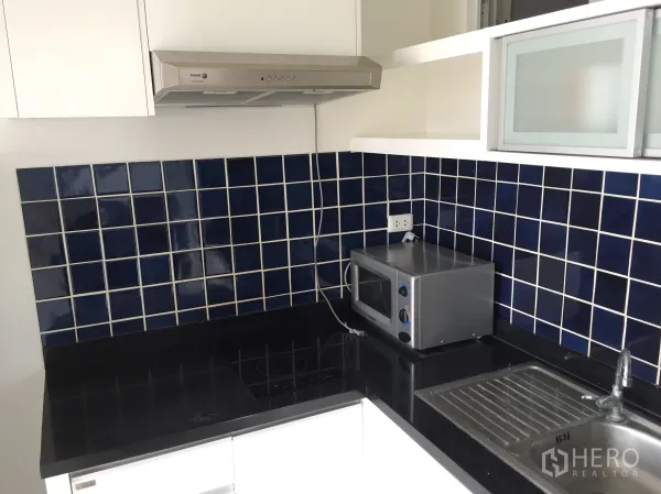 Condominium for rent in Khlong Toei, Bangkok - Separate kitchen with dark countertops, blue tiled backsplash, hob, hood, microwave and sink.