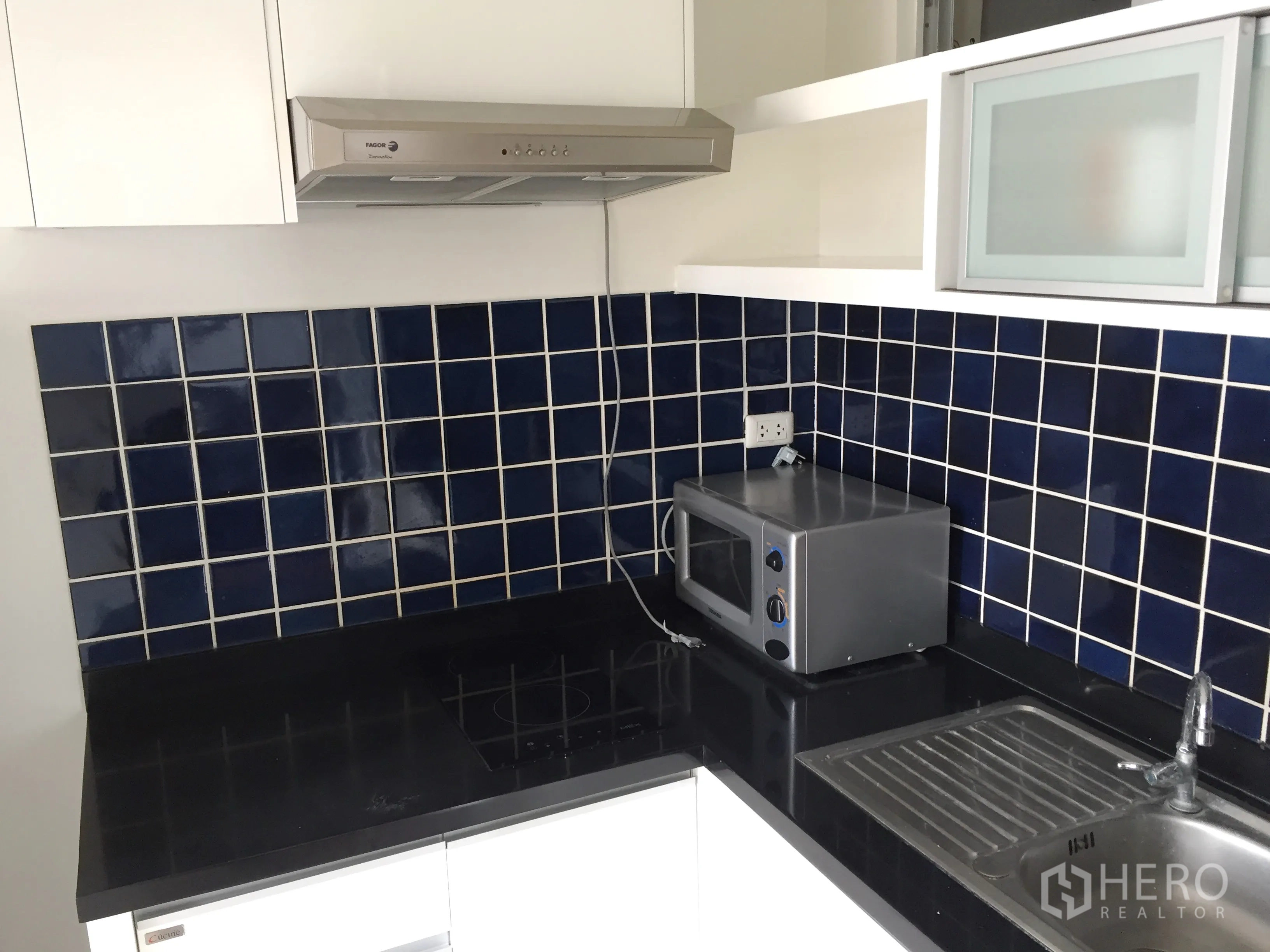 Condominium for rent in Khlong Toei, Bangkok - Separate kitchen with dark countertops, blue tiled backsplash, hob, hood, microwave and sink.