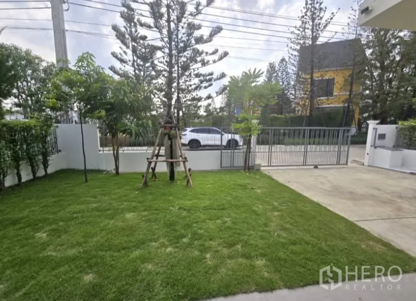 独立式住宅 for rent in Bang Phli, Samut Prakan - 前庭草坪与车道，金属大门及乔木点缀。