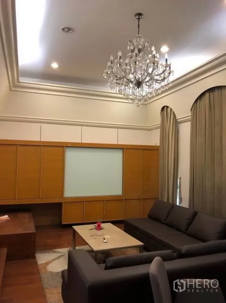 Townhouse for rent in Chatuchak, Bangkok - Living room with crystal chandelier, L-shaped sofa, wood cabinets and tall draped windows.