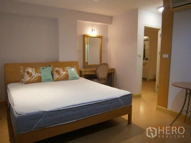 Condominium for rent in Khlong Toei, Bangkok - Bright master bedroom with wooden bed, vanity desk with mirror, and a doorway leading to the bathroom.