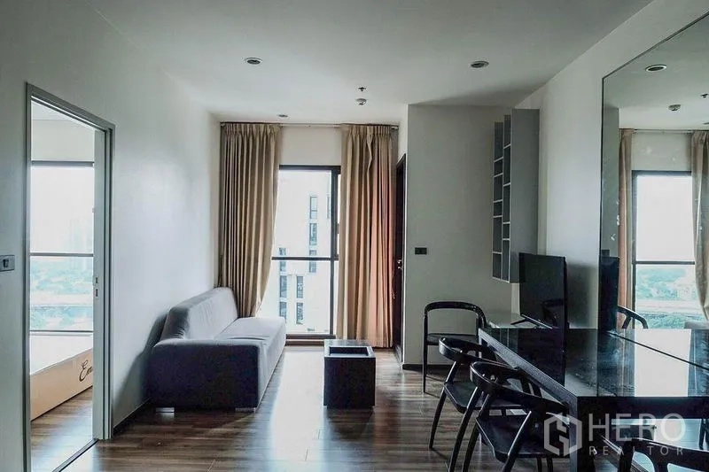 Condominium for rent in Khlong Toei, Bangkok - Open living and dining space with sofa, tall windows and TV shelving.