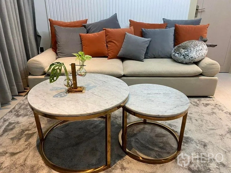 Condominium for sale in Huai Khwang, Bangkok - Cozy living area with cream sofa, orange and gray cushions, and round marble nesting coffee tables with gold frames.