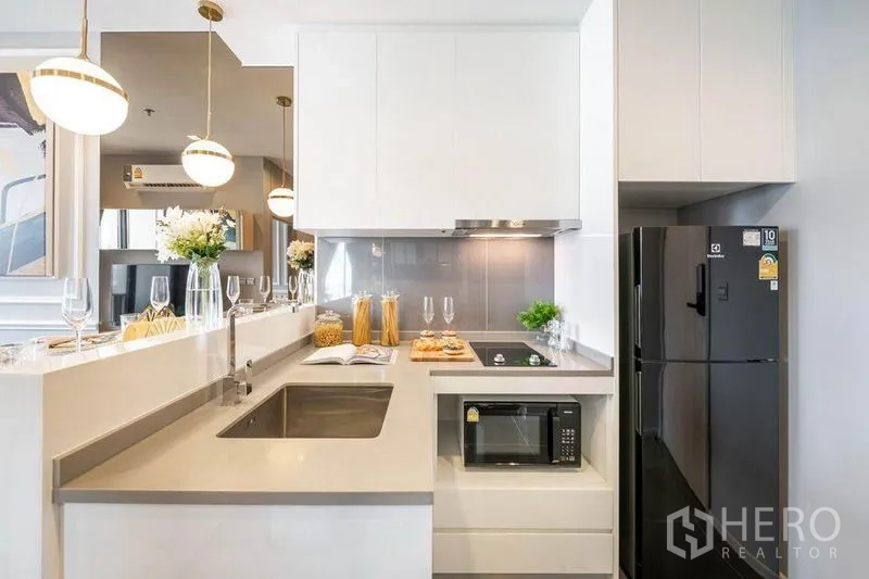 Condominium for rent or sale in Watthana, Bangkok - Modern white kitchen with breakfast bar, sink, cooktop, microwave and a black fridge.